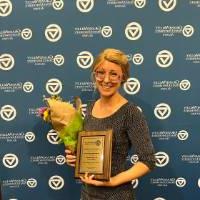 Alum standing in front of the GV Alumni Relations backdrop with an award and flowers.